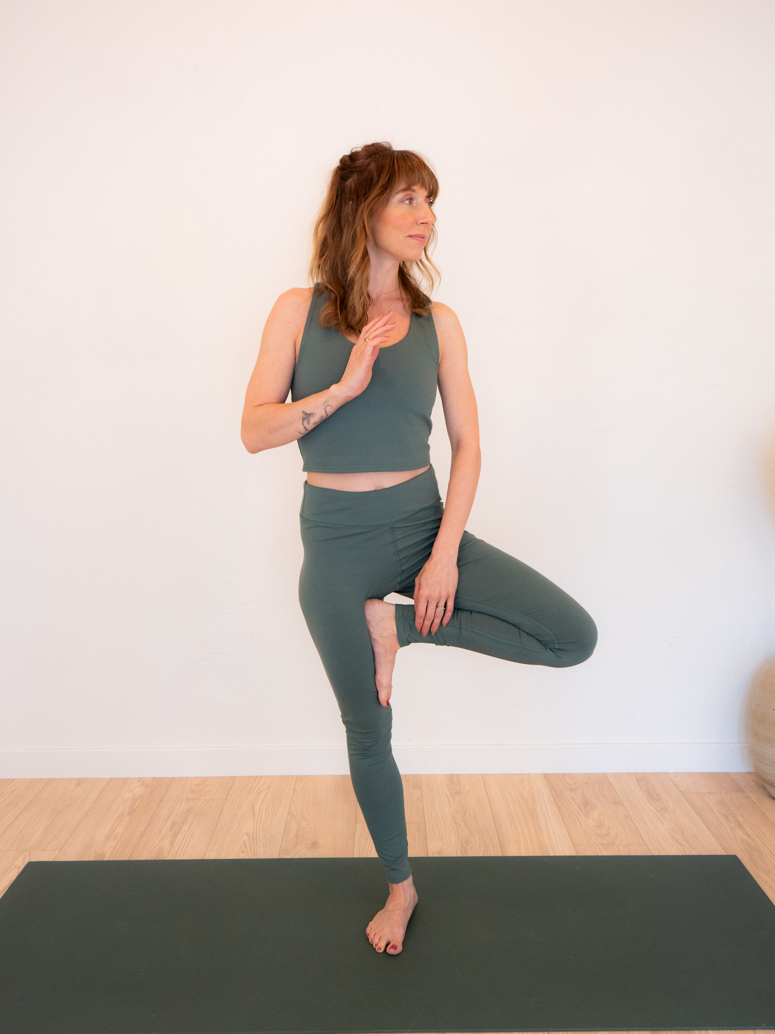 A person performs a yoga tree pose on a mat, wearing a green sleeveless top and leggings. The background is a plain white wall.
