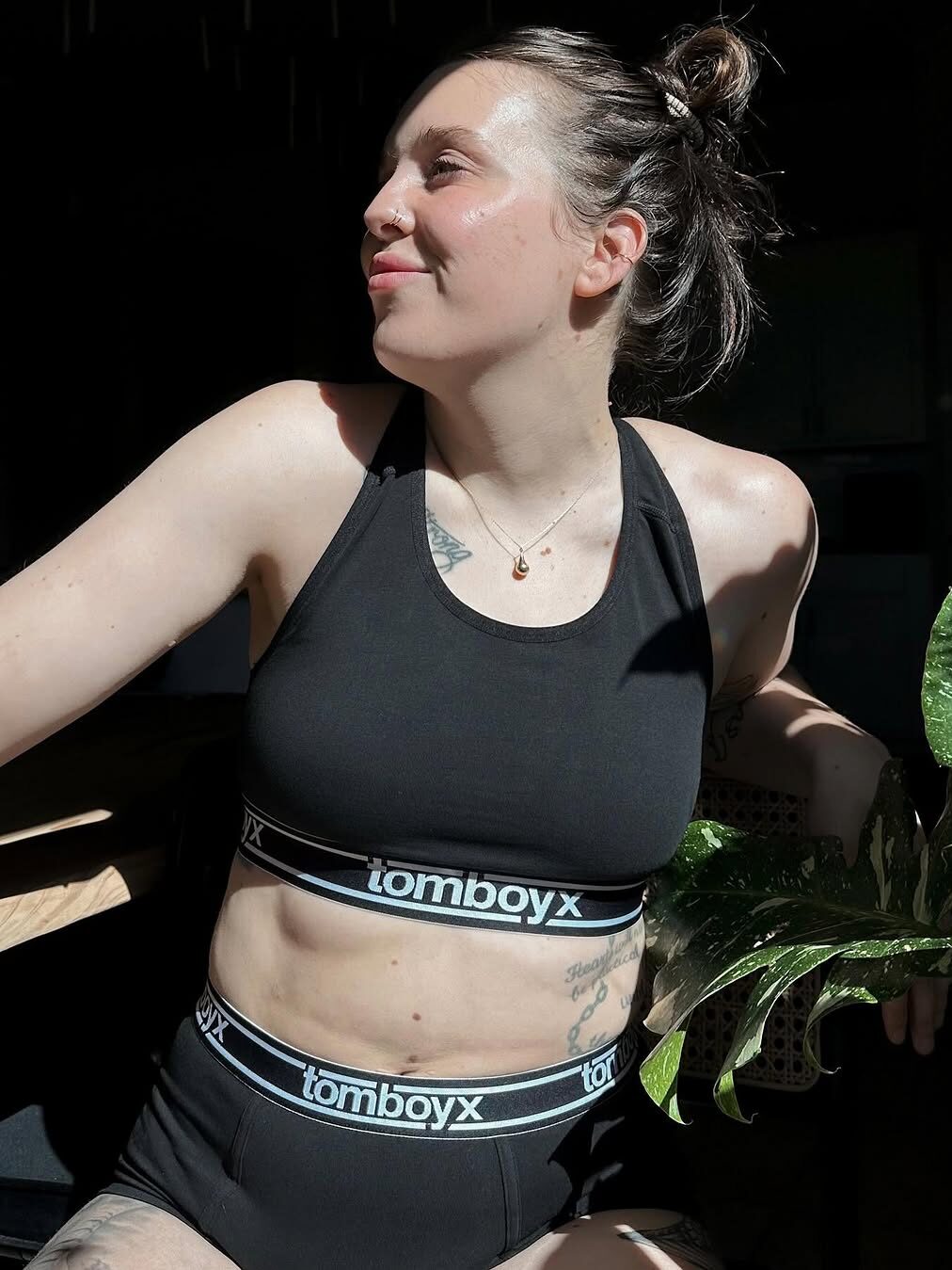Person in black athletic wear sits with a plant, looking to the side, in a sunlit setting.