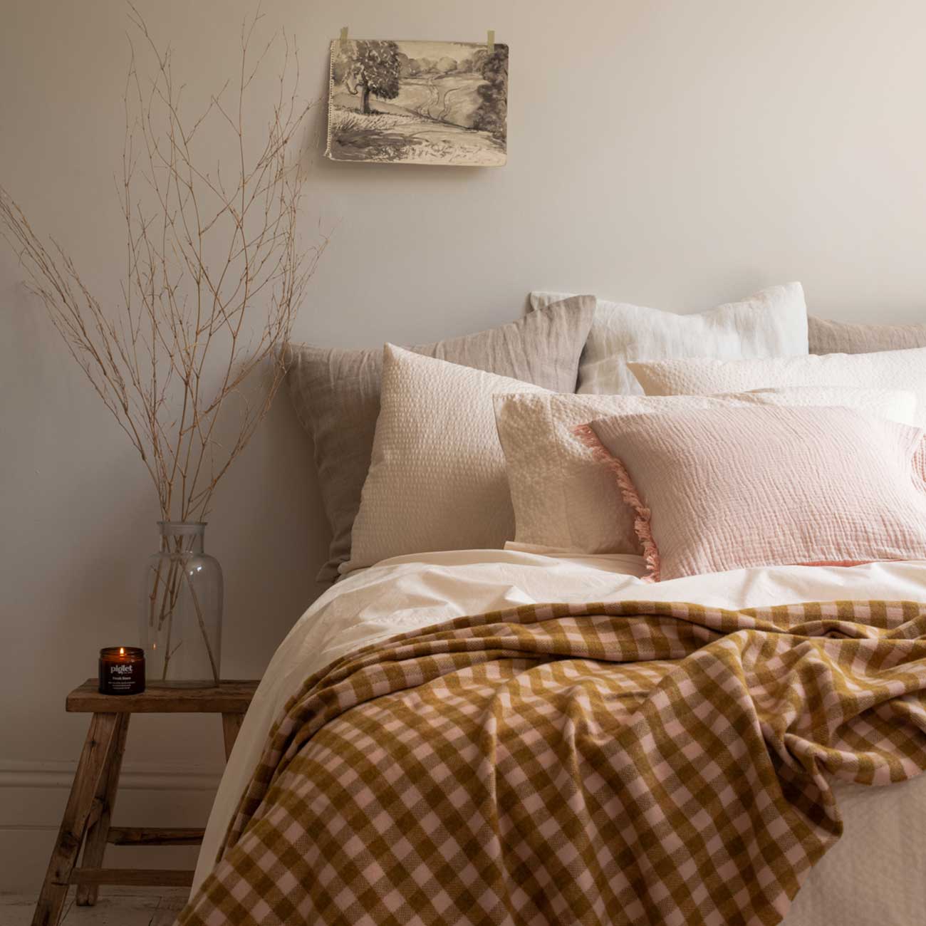 A neatly made bed with neutral-toned pillows and a brown gingham blanket, a wooden stool with a vase and candle, and a small artwork hanging on a pale wall.