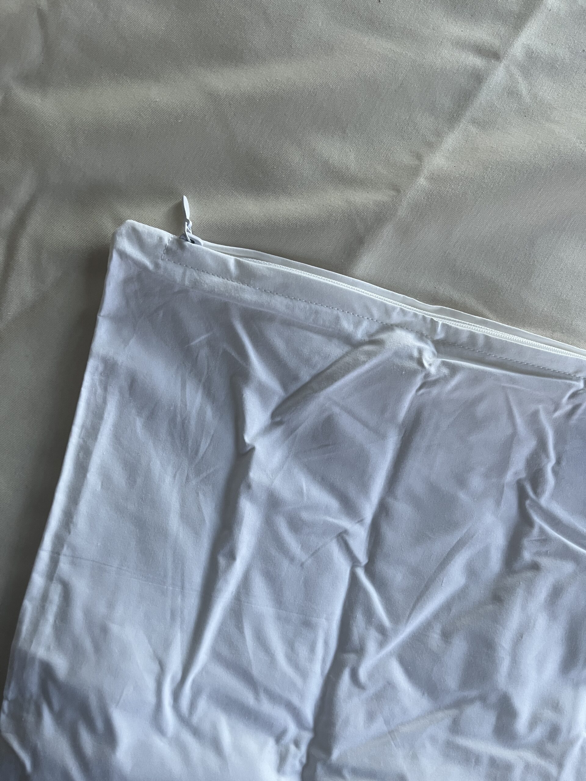 White, wrinkled zippered pillowcase partially shown on a similar-colored fabric background.