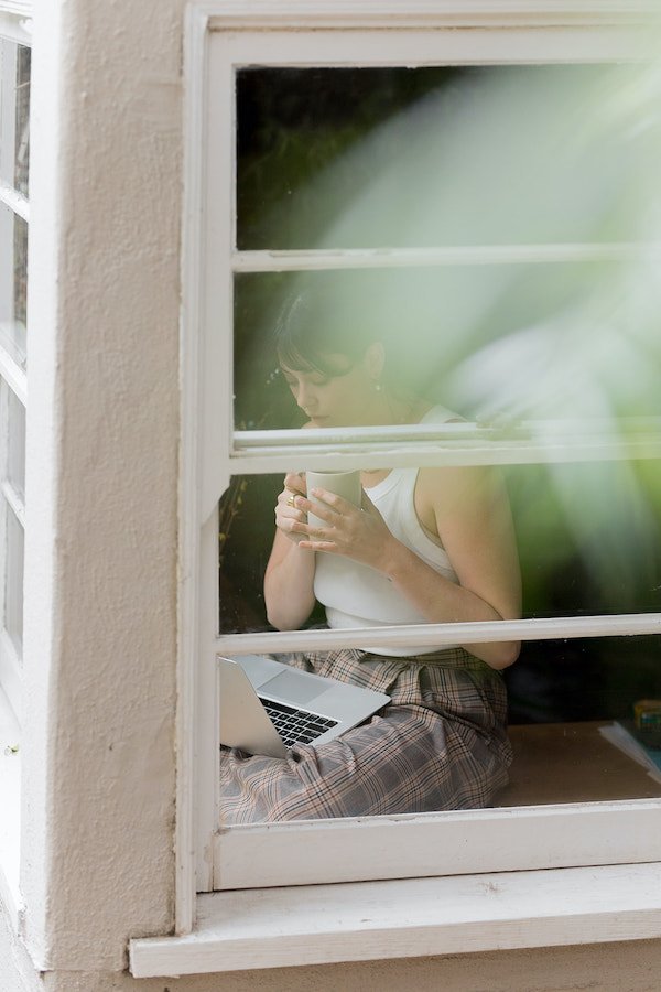 Person sitting by a window, drinking from a cup, with a laptop on their lap. A plant is partially visible outside the window.