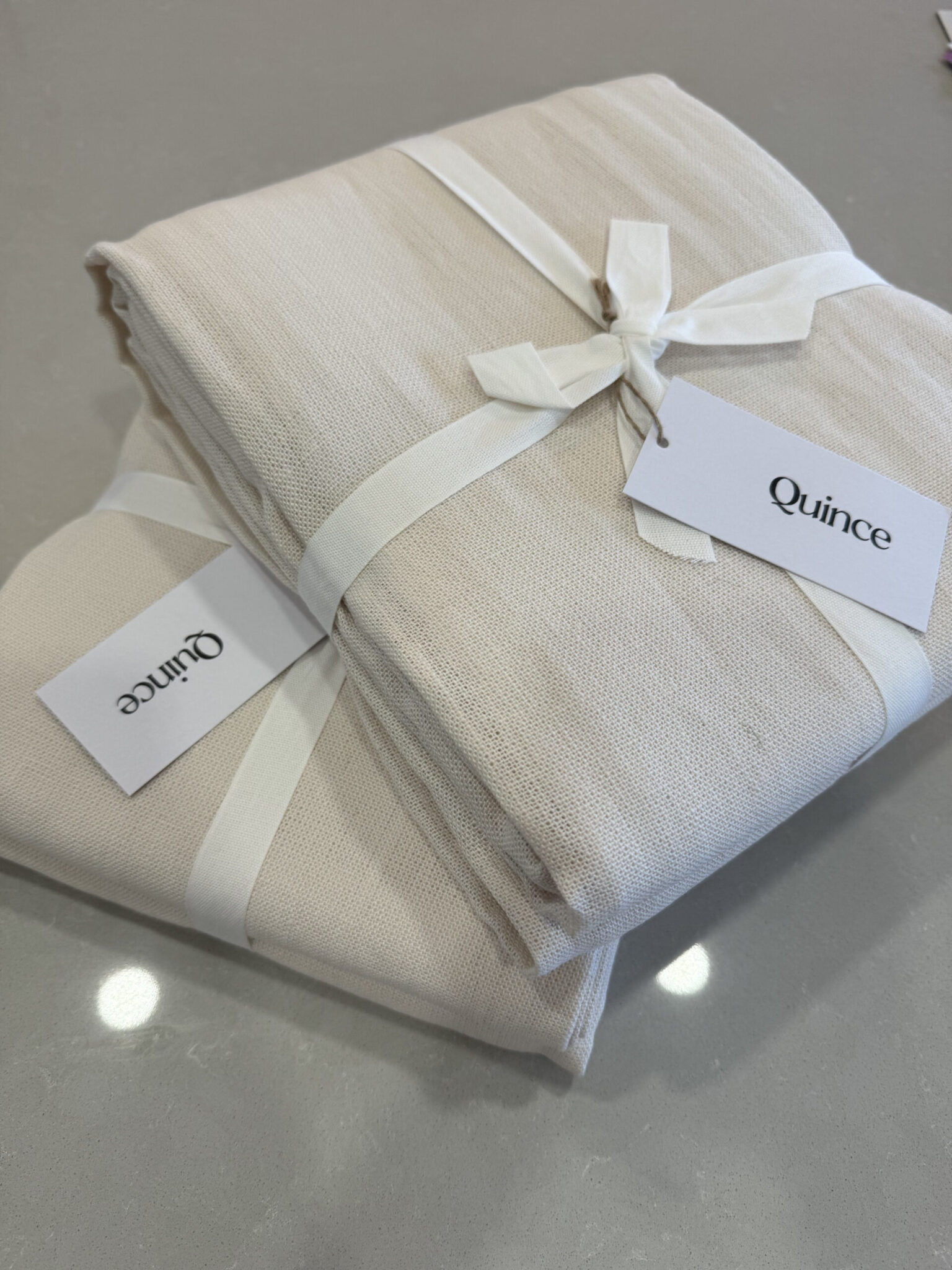 Two folded beige fabric bundles tied with white ribbons, each with a Quince brand tag, placed on a smooth gray surface.
