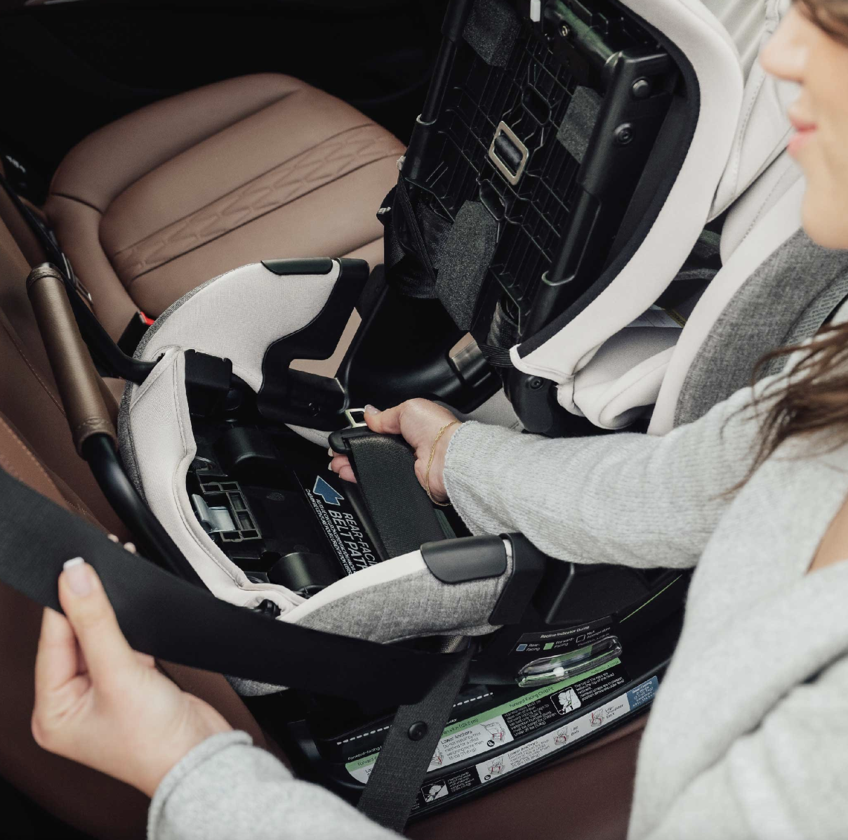Person adjusting a black seatbelt on a gray infant car seat inside a car with brown leather seats.