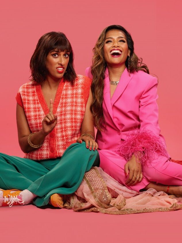 Two women sitting on the floor with a pink background. One wears a red checkered top and green pants, the other a pink suit. Both are smiling.