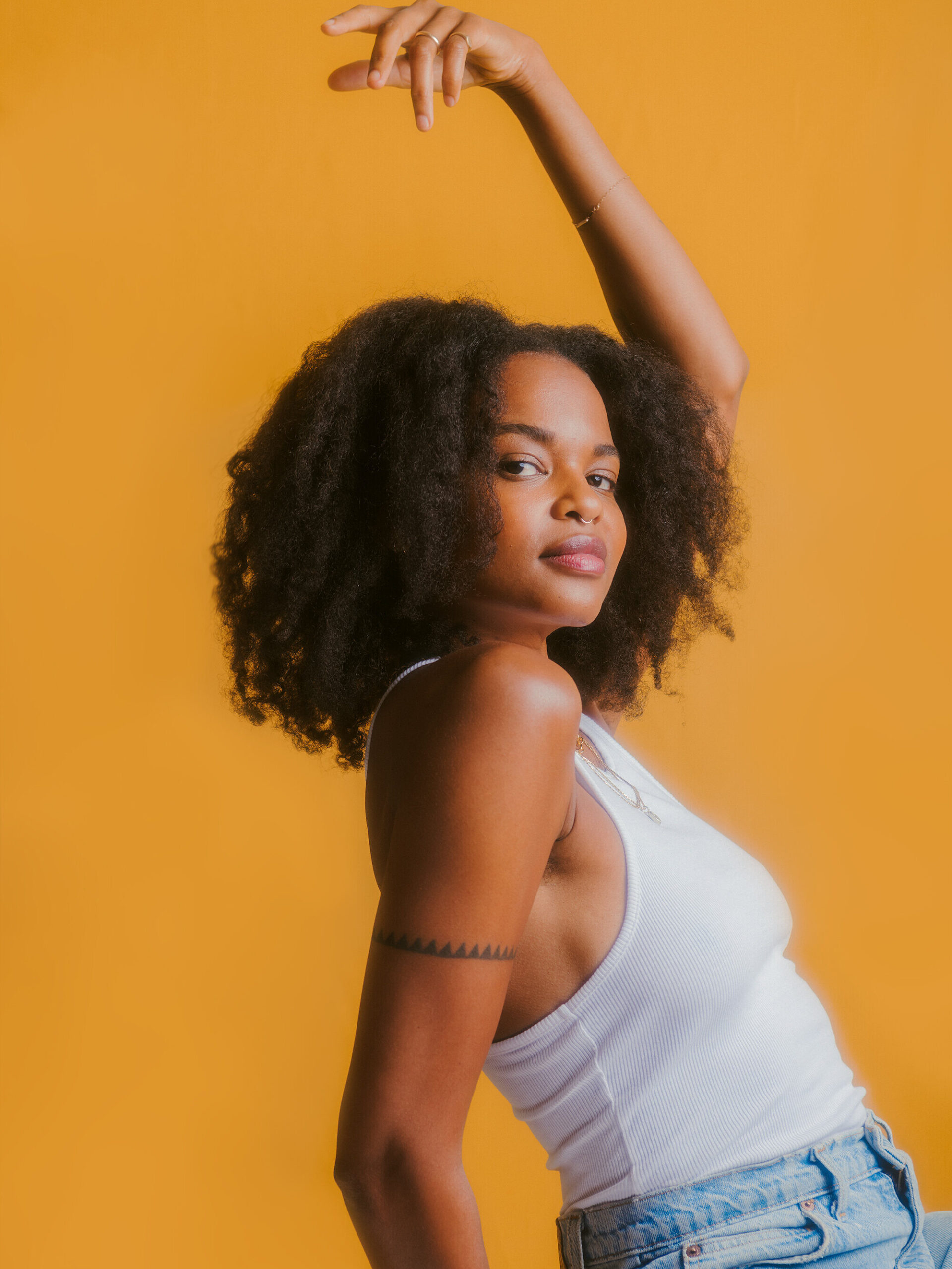 Person with curly hair wearing a white tank top and jeans poses against an orange background with one arm raised.