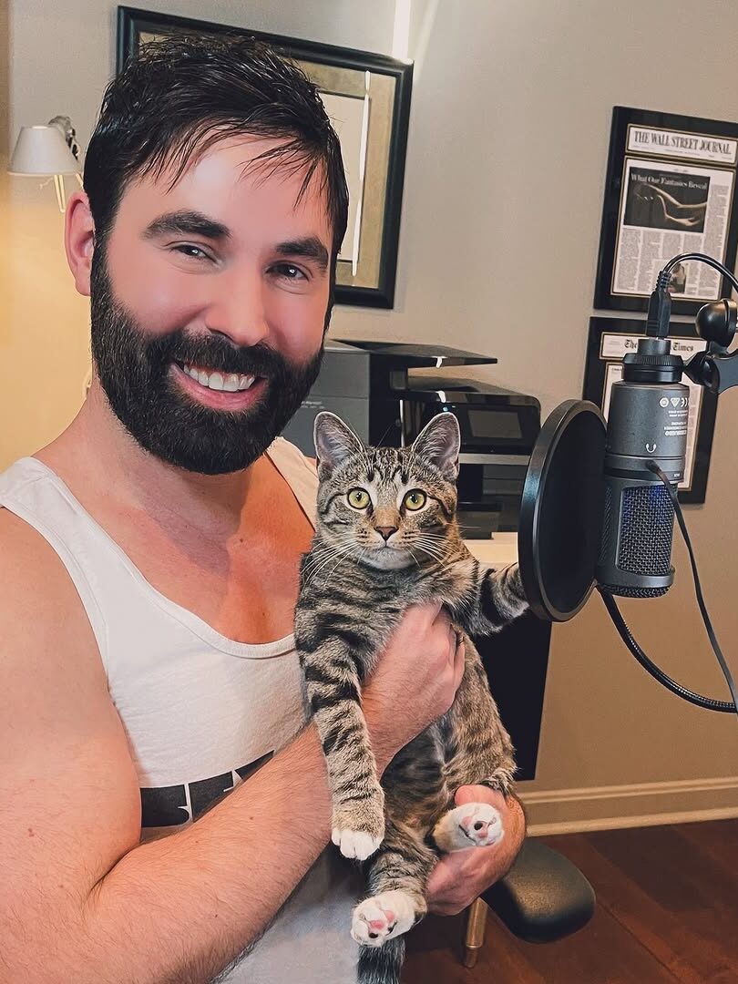 Person smiling while holding a tabby cat in front of a microphone, seated in a home office with framed newspapers on the wall.