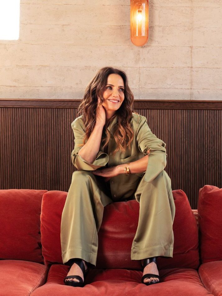 Person sitting on a red couch, wearing a green outfit, with a calm expression, in a warmly lit room with a wooden wall and light fixture.