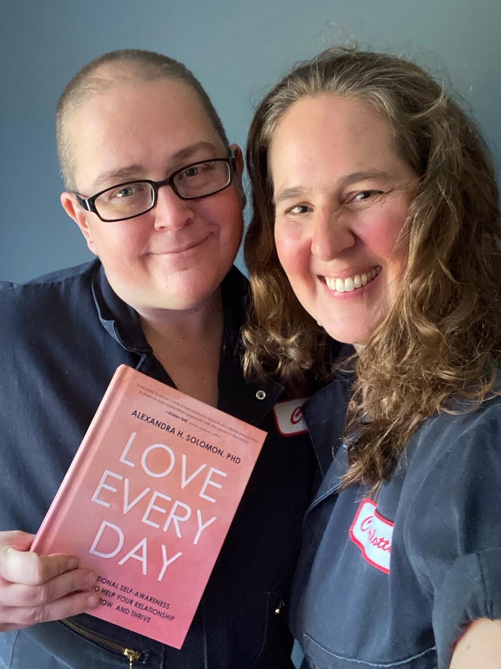 Two people wearing matching dark jackets smile. One holds a book titled "Love Every Day.