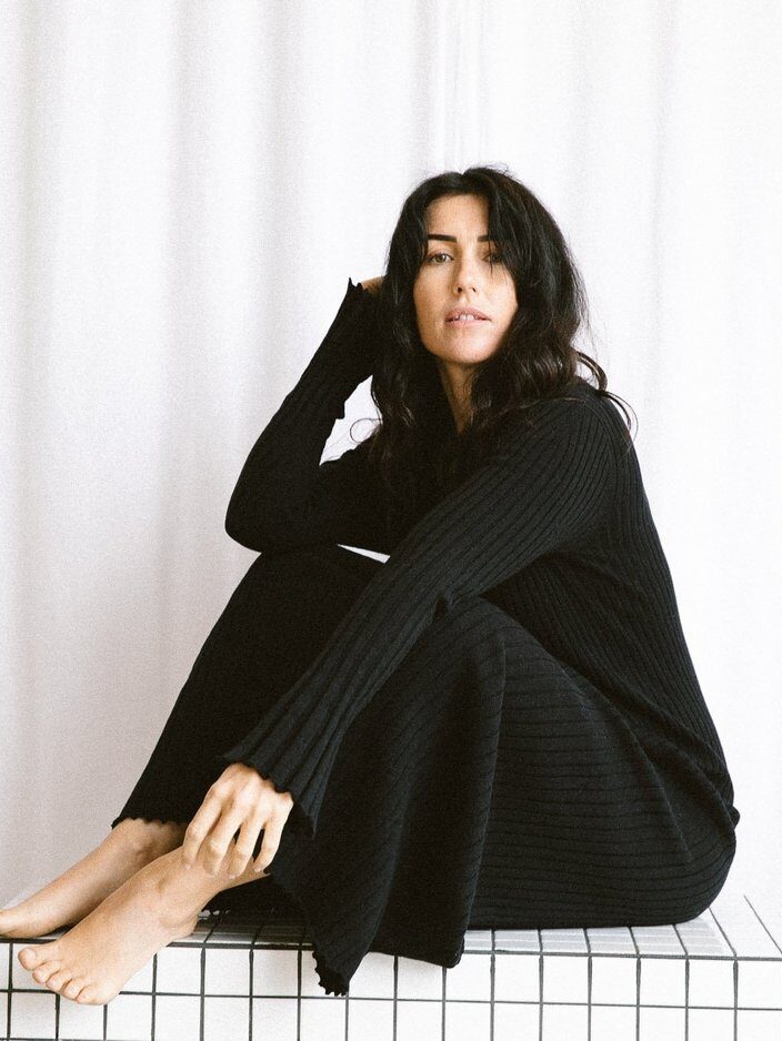 Person sitting with legs folded, wearing a black ribbed outfit, in front of a white curtain.