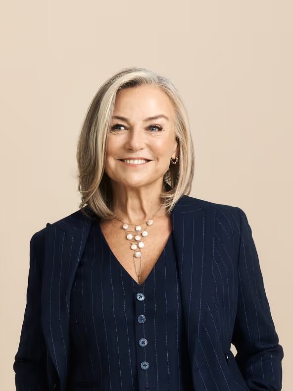 Smiling person with gray hair wearing a dark pinstripe suit and a pearl necklace, standing against a beige background.