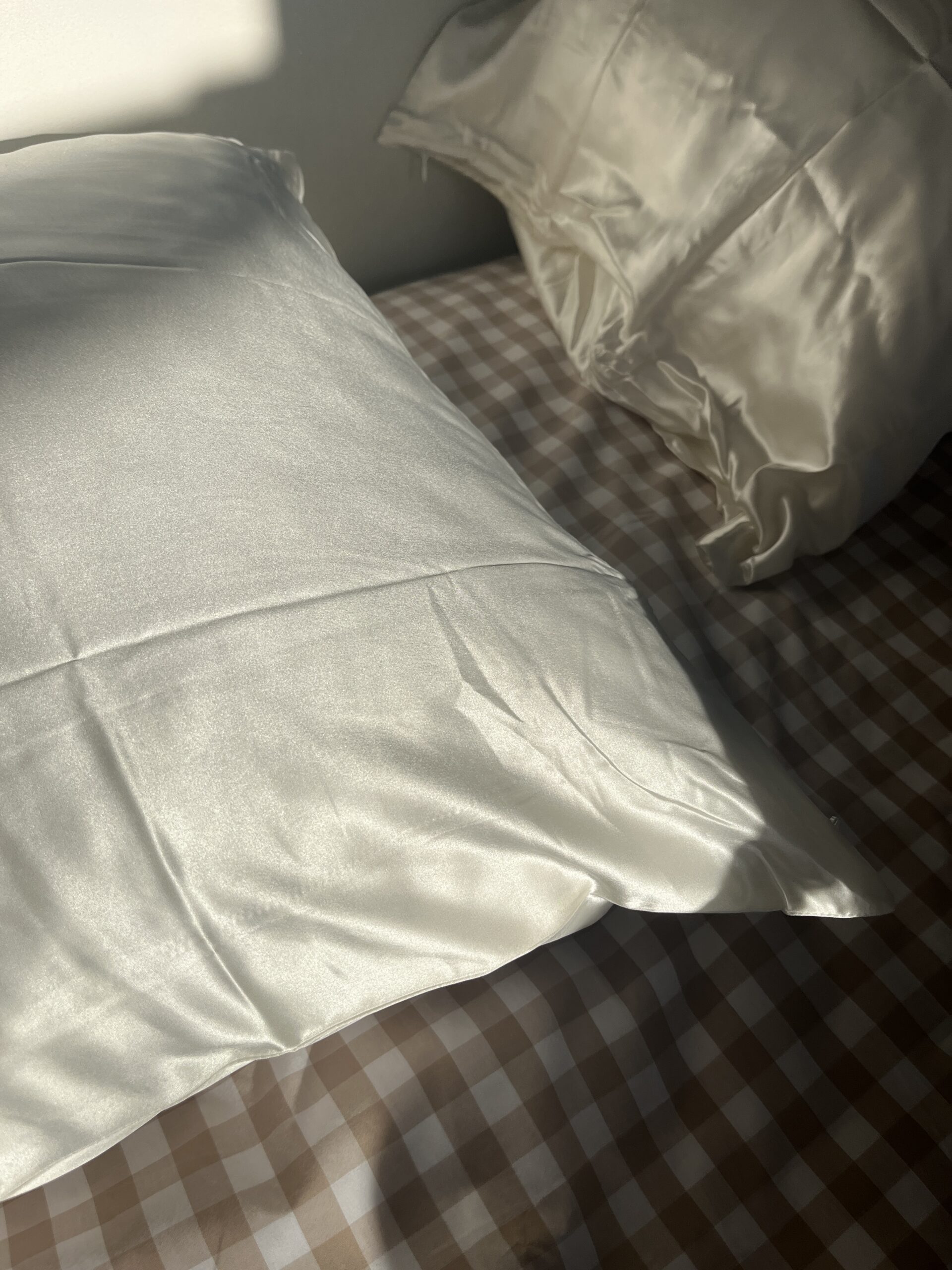 Two white satin pillowcases on a bed with a brown and white checkered sheet, partially illuminated by sunlight.