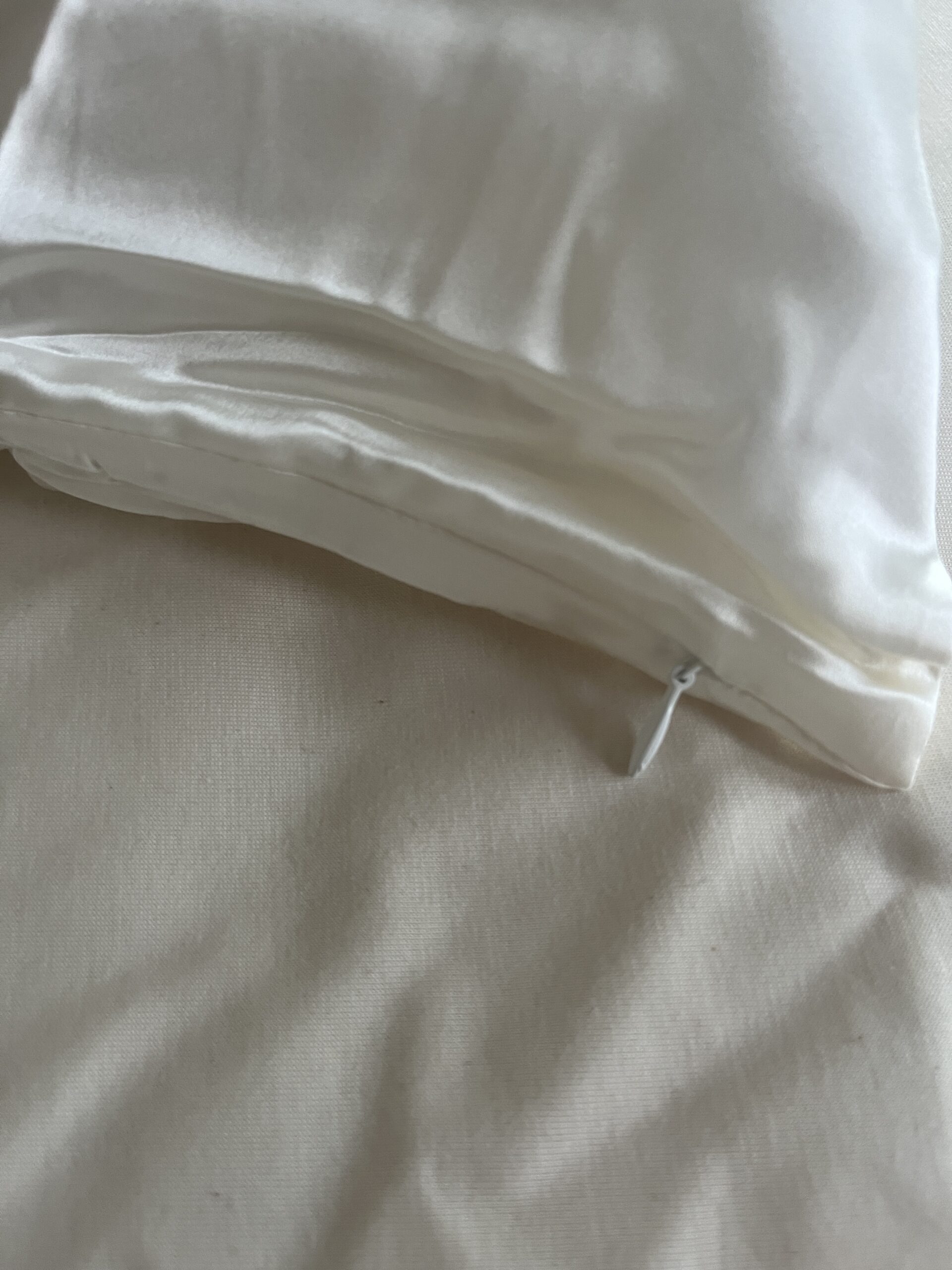 Close-up of a white silk pillowcase with a visible zipper on a cream colored surface.