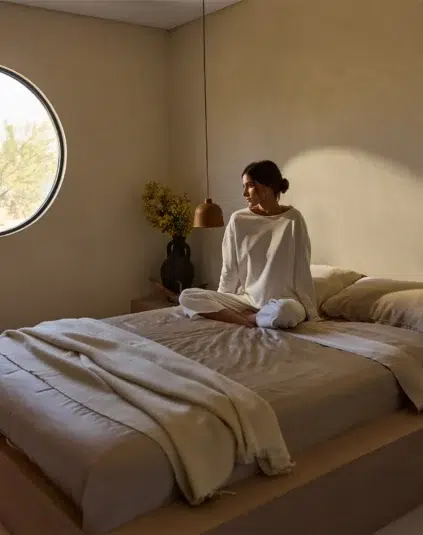 A woman in white loungewear sits cross-legged on a neatly made bed in a minimal, sunlit bedroom with beige walls and a round window.