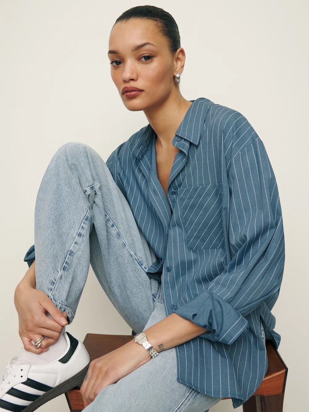 Person sitting on a stool wearing a blue pinstripe shirt, light jeans, and white sneakers, with hair tied back and neutral expression.