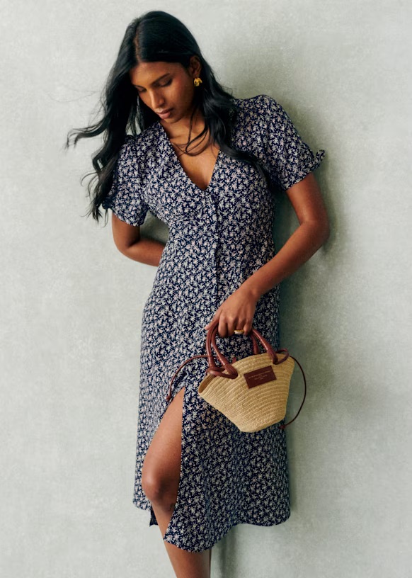 Person wearing a floral dress holding a straw handbag, standing against a neutral background.