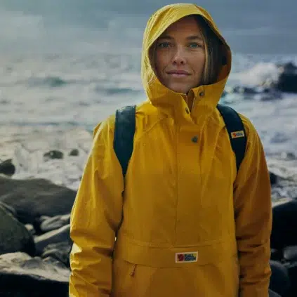 A person in a waterproof yellow raincoat stands by a rocky shoreline, waves crashing in the background. They wear a hood and carry a backpack, ready for adventure.