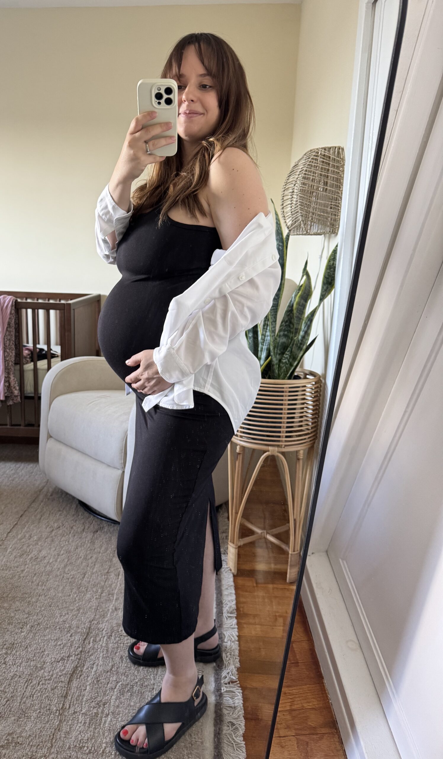 A person in a black dress and white shirt takes a side mirror selfie, showing a pregnancy bump. There is a crib, armchair, and plant in the background.
