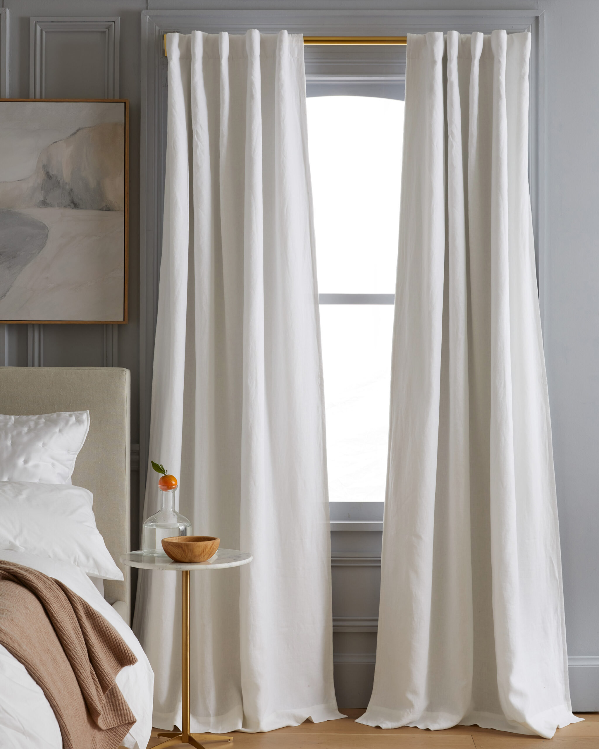 White curtains hang in front of a window in a room with a small round table, a bed with white bedding, and a wall painting.