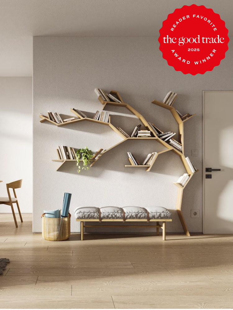 Tree-shaped wooden bookshelf on a white wall, holding books. Below, a cushioned bench with an umbrella and a basket beside it. "The Good Trade 2023 Reader Favorite Award Winner" badge in the corner.