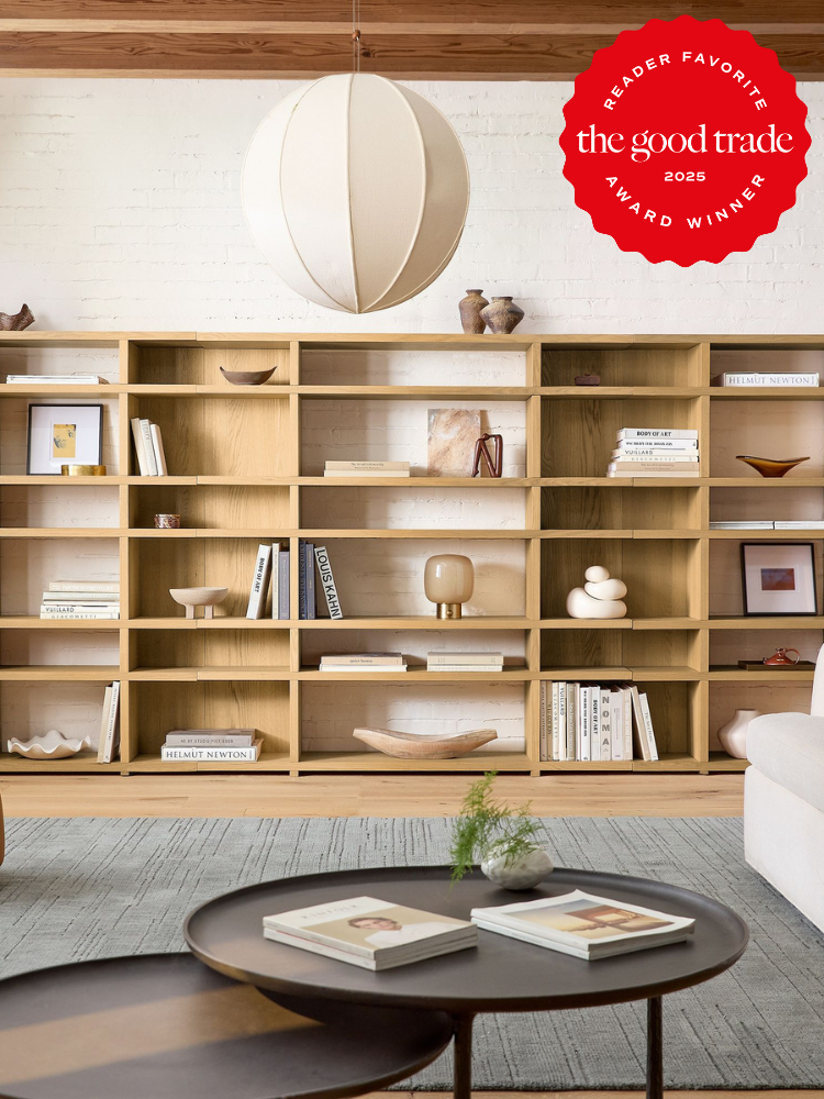 Modern living room with a large wooden bookshelf, decorative items, a white sofa, and round coffee tables. A "Reader Favorite" award from "The Good Trade" is displayed in the image.