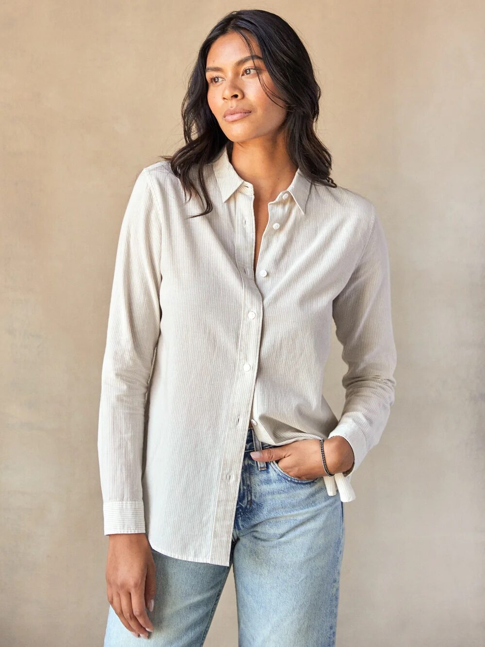 A person wearing a light beige button-up shirt and blue jeans stands against a neutral background, looking to the side with a relaxed expression.