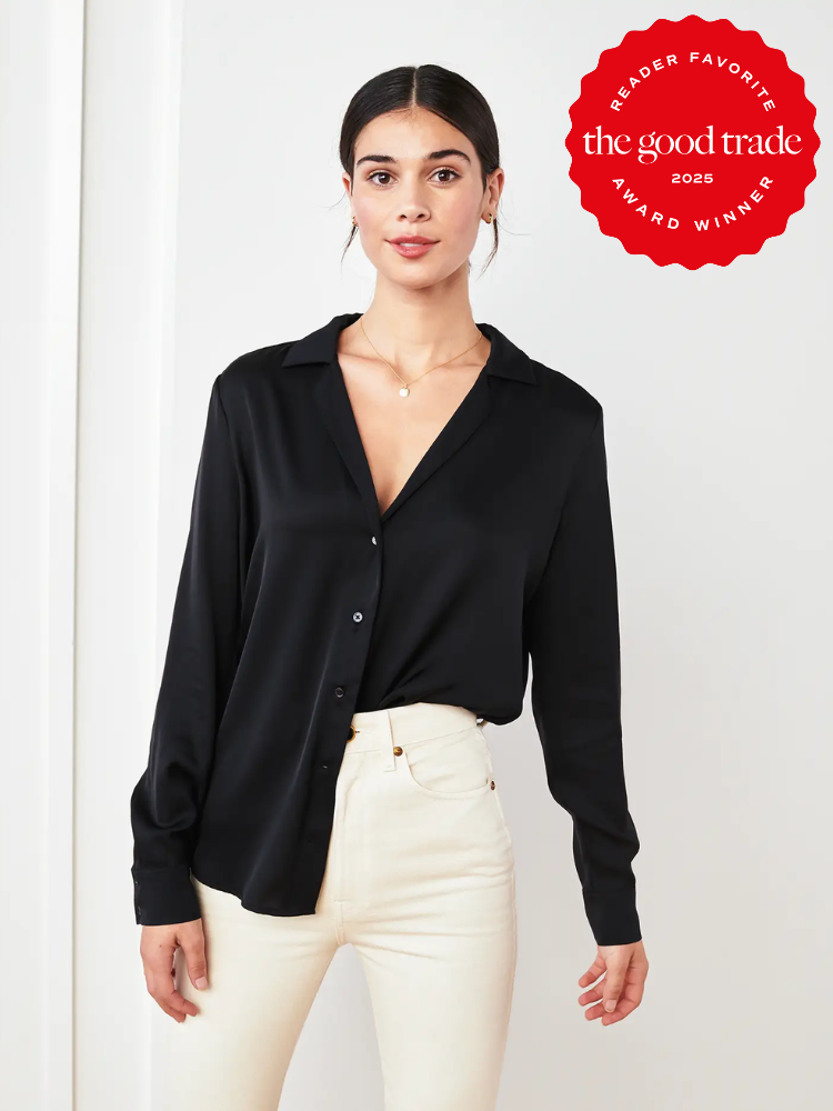 Person wearing a black button-up shirt and white pants stands indoors near a wall. A red "The Good Trade" award emblem is in the top right corner.