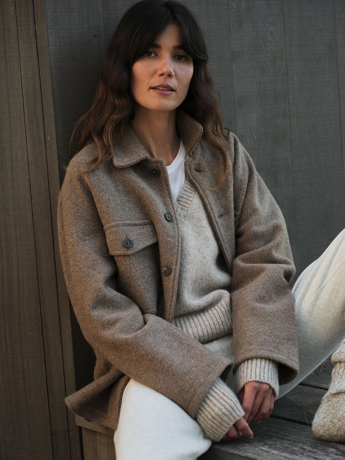 A person sits against a wooden wall wearing a grey coat, light sweater, and white pants. They have long brown hair and a relaxed expression.