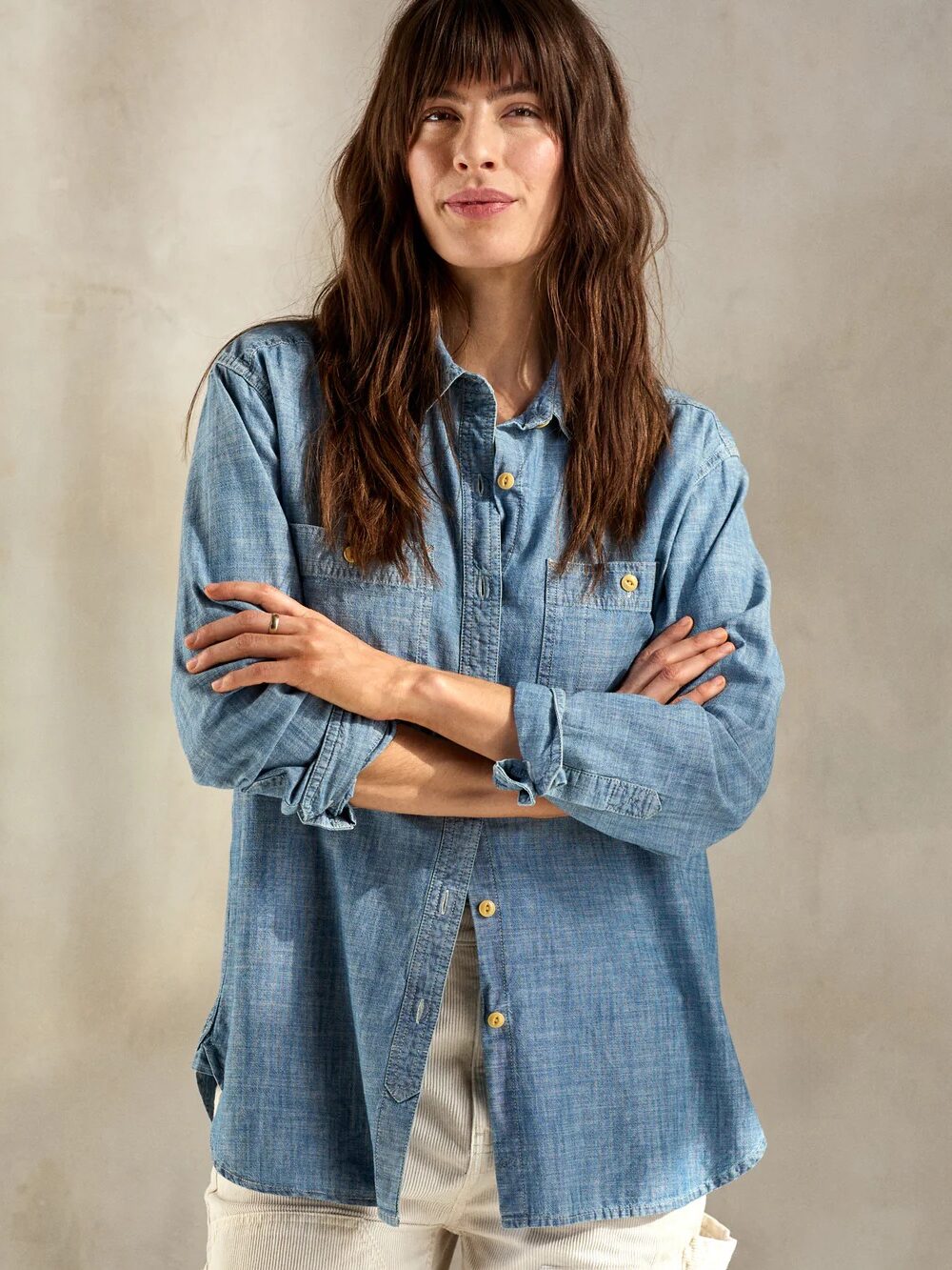 A person with long hair stands with arms crossed, wearing a blue denim shirt and light-colored pants, against a neutral background.