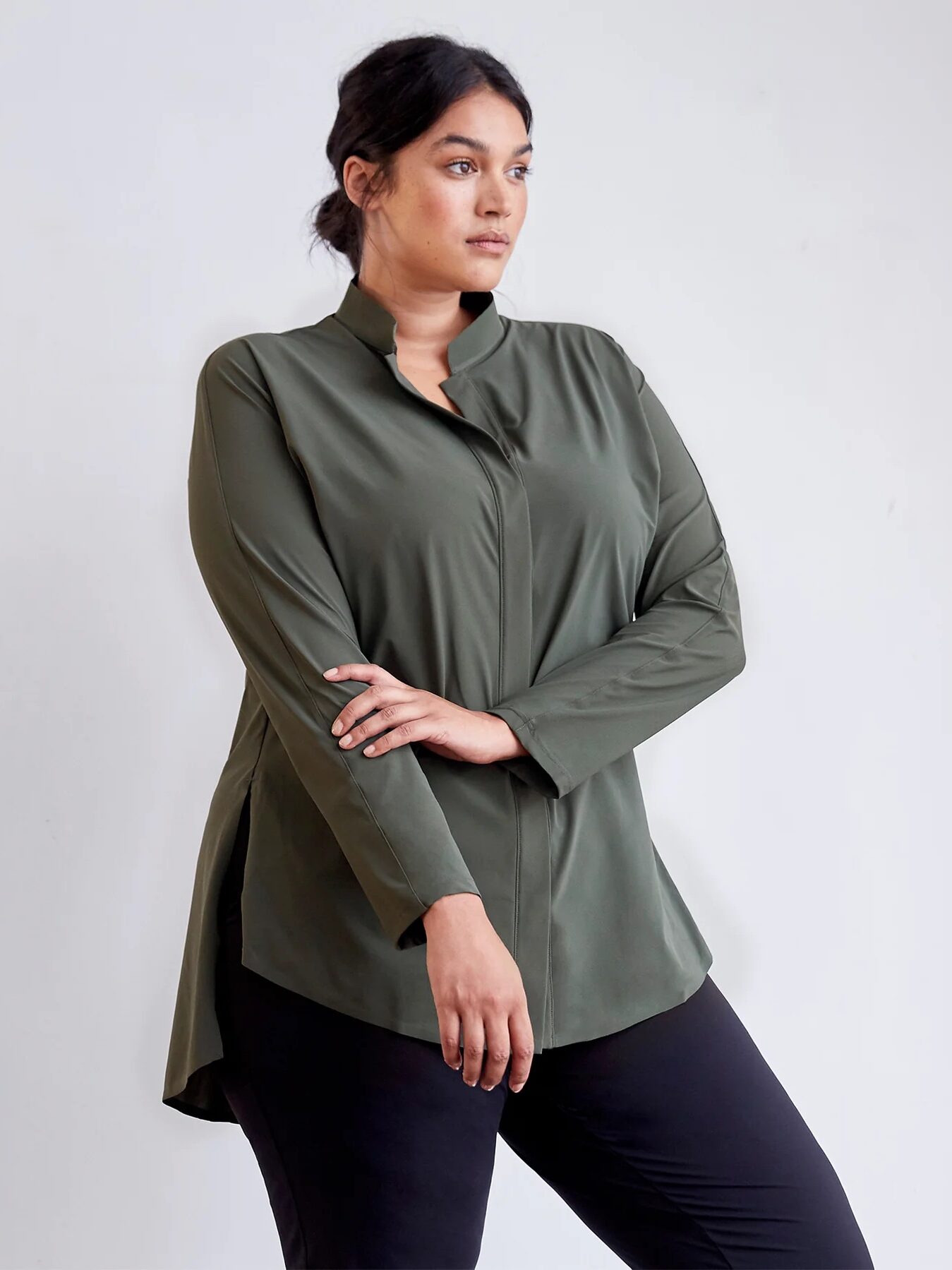 Person wearing a long-sleeved, olive green blouse with hands crossed, paired with black pants, against a plain background.