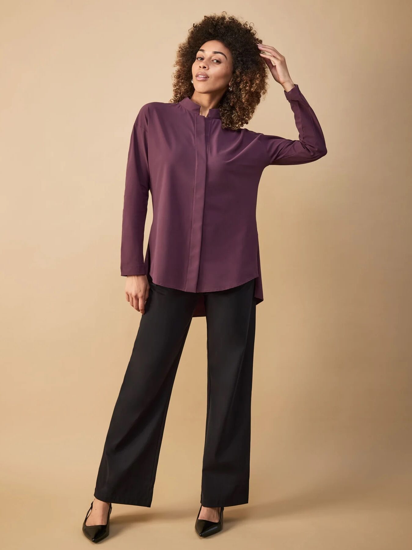 Woman wearing a purple shirt and black pants, standing against a beige background.