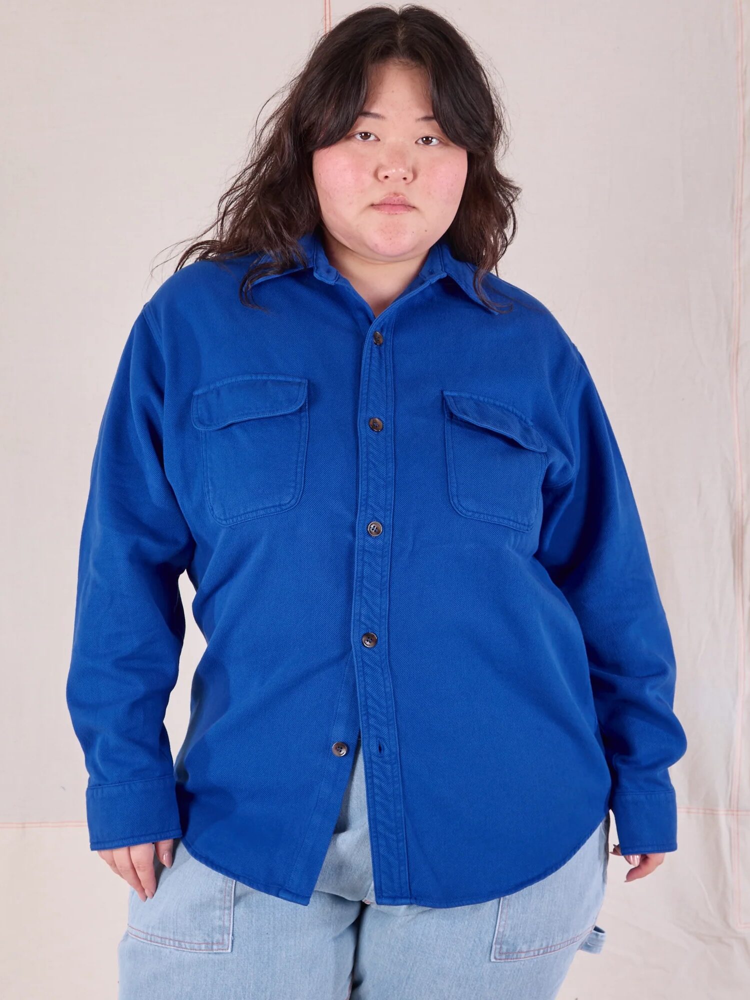 A person with long hair is wearing a blue button-up shirt with two chest pockets and light blue jeans, standing against a beige backdrop.