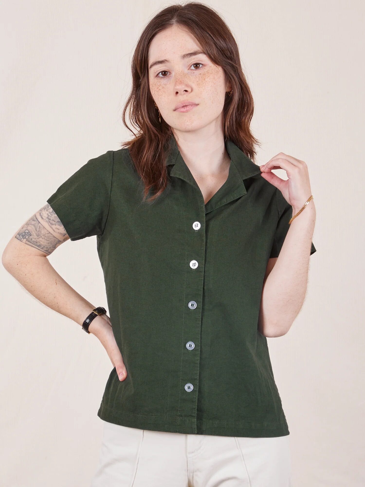 Person wearing a dark green button-up shirt and white pants poses against a plain background.