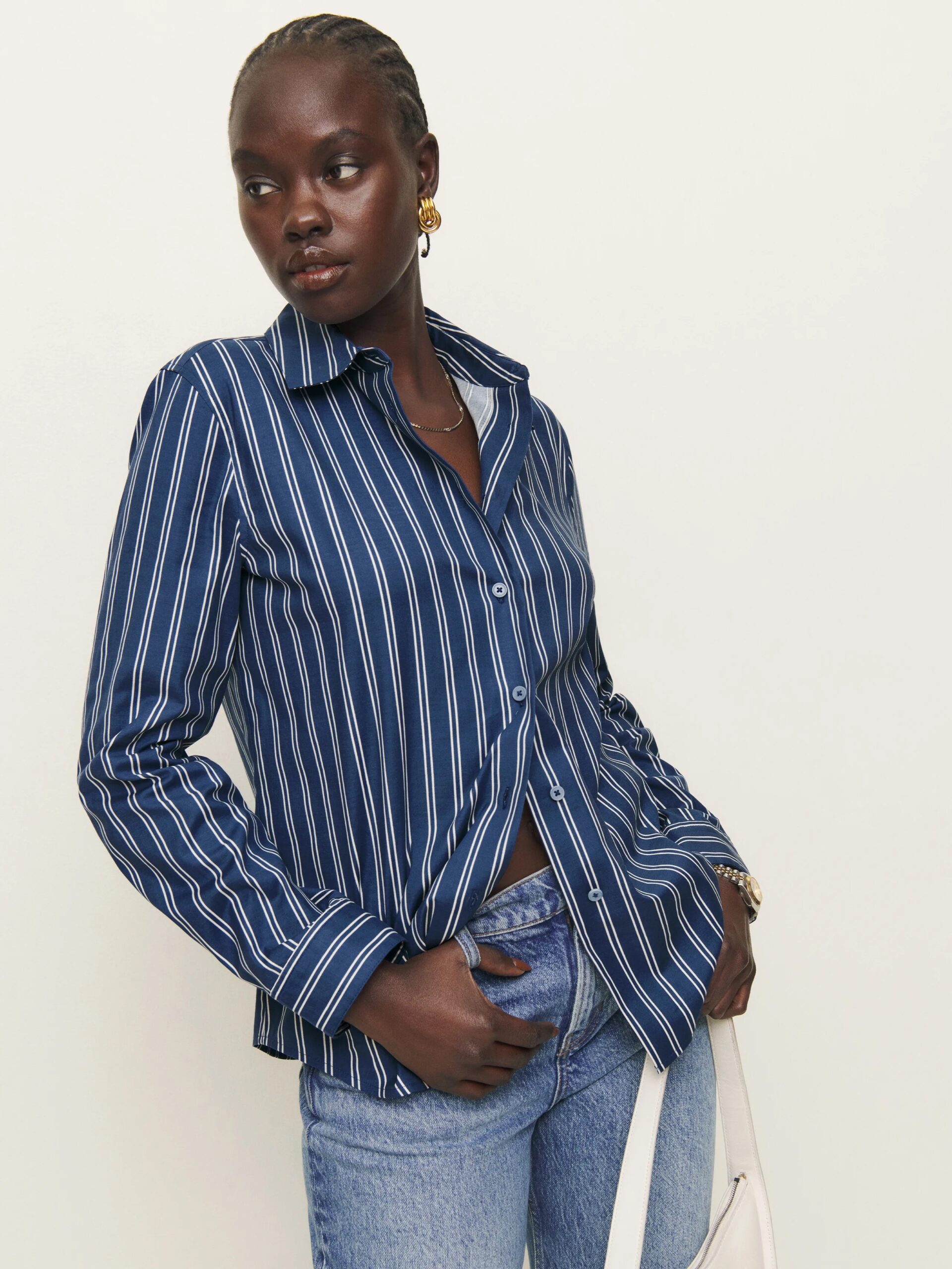 A person in a blue and white striped shirt and jeans, standing against a plain background. They have their hand in their pocket and are looking to the side.