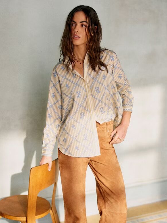 Person with long hair wearing a patterned shirt and brown pants, standing beside a wooden chair.