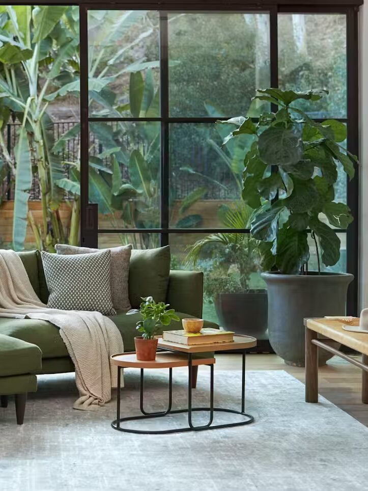 A modern living room with a green sectional sofa, green and gray cushions, two wooden tables, and large plants. A glass wall reveals lush greenery outside.