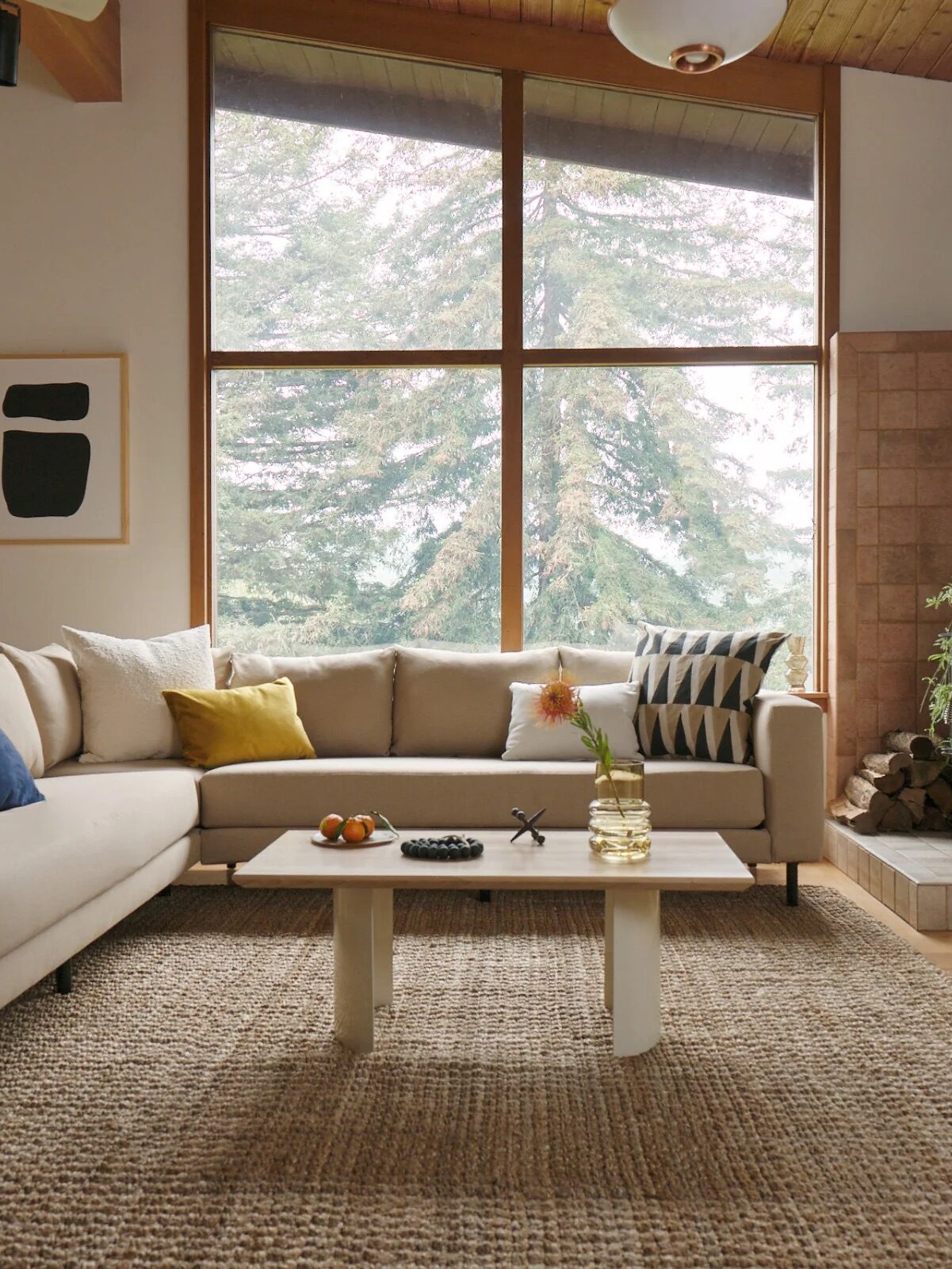 Cozy living room with a beige sectional sofa, a small coffee table, and a wood-burning stove. Large windows showcase a view of trees. Minimalist decor includes abstract art and plants.