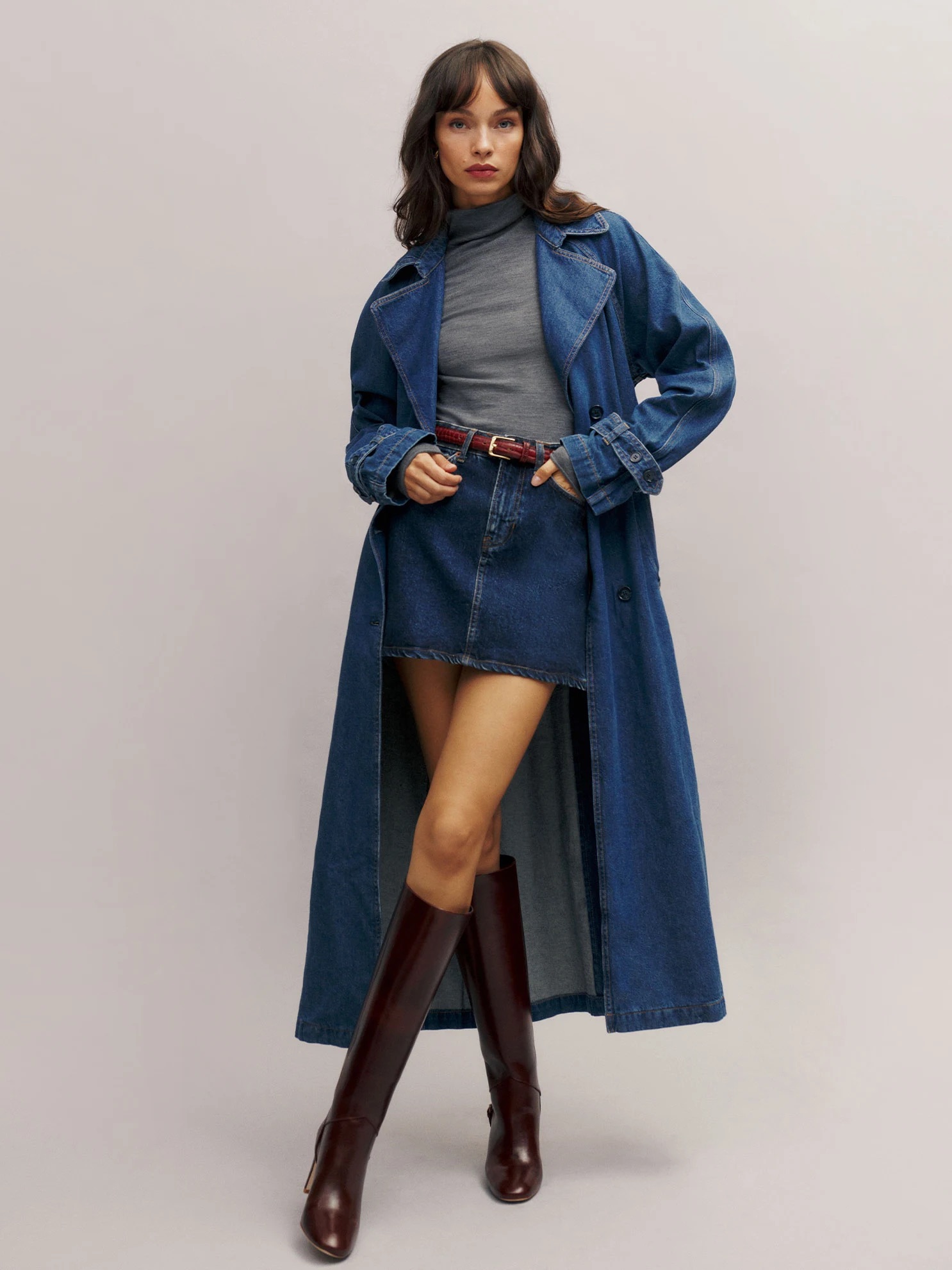 A person wearing a long denim coat, gray turtleneck, denim skirt, and brown knee-high boots stands against a plain backdrop.