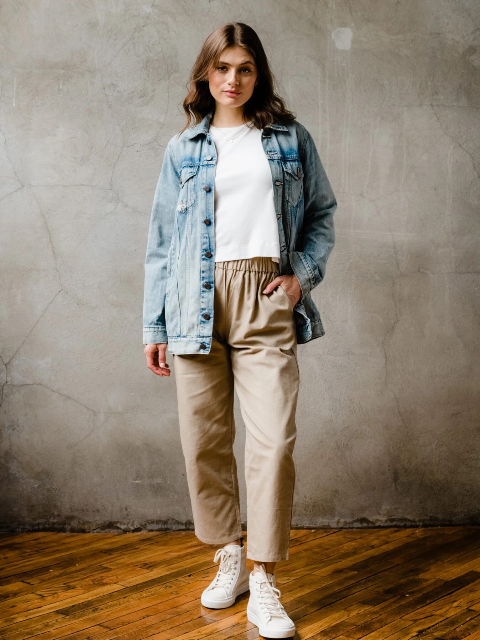Person in a denim jacket, white shirt, tan pants, and white sneakers stands on a wooden floor against a gray wall.