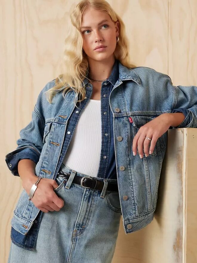 Person with long blonde hair wearing a denim jacket over a denim shirt and skirt, leaning against a wooden surface.