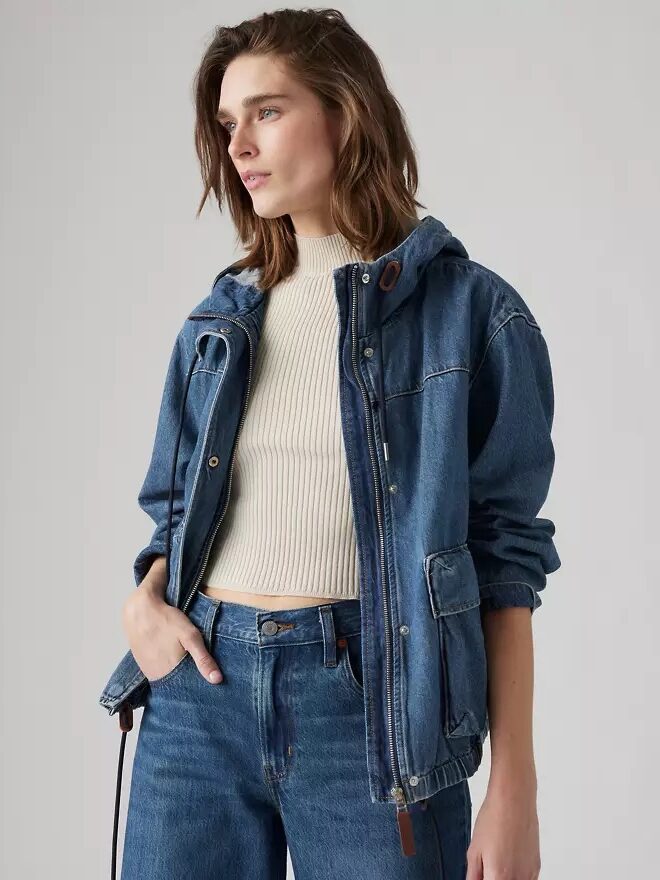 Person wearing a denim jacket and jeans, with hands in pockets, looking to the side against a plain background.