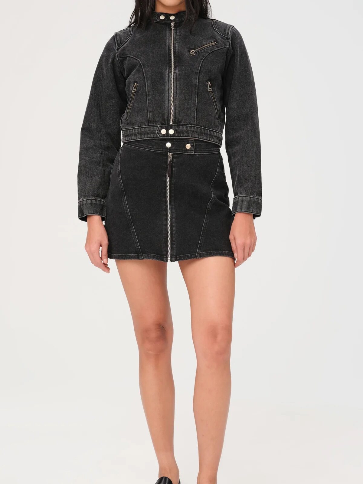 Person wearing a black denim jacket and skirt with zipper details, paired with black loafers, stands against a plain white background.