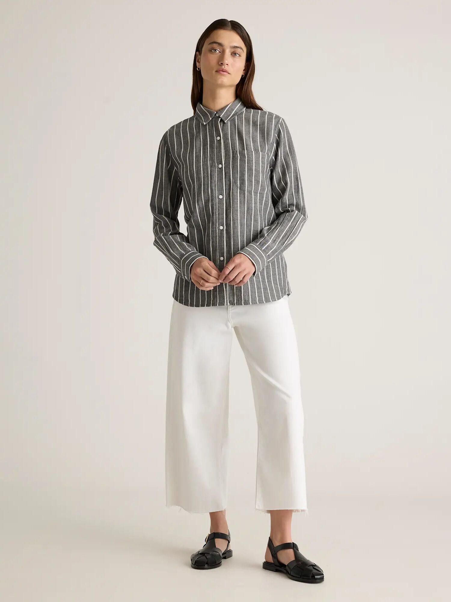 A person wearing a dark striped shirt and white wide-leg pants stands against a plain background.