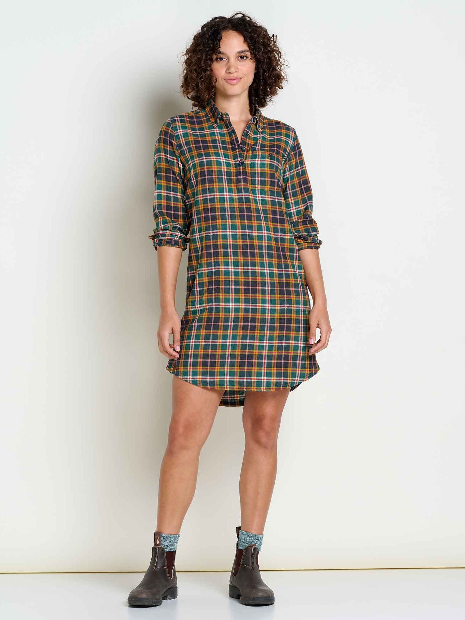 Person wearing a plaid long-sleeve shirt dress, with rolled-up sleeves, standing against a plain background, wearing boots and socks.