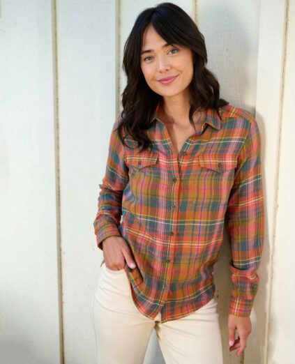 A person with long dark hair wearing a multicolored plaid shirt and light pants smiles while leaning against a wall.