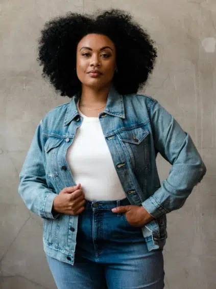 A person with curly hair wearing a denim jacket and jeans stands against a textured wall, facing forward.
