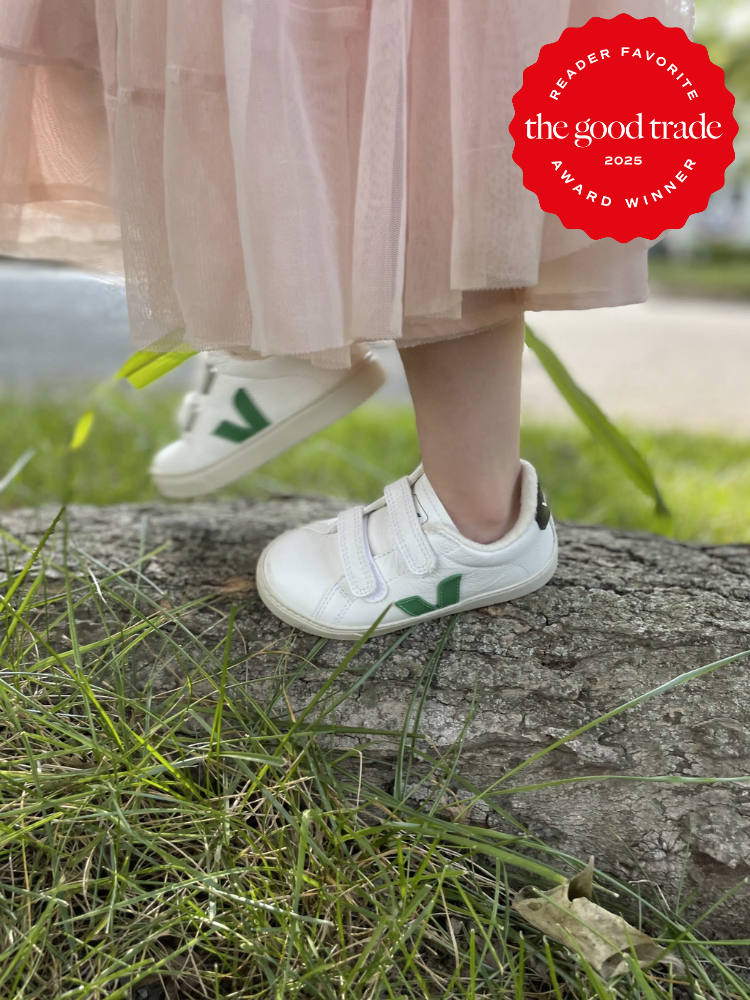 Child wearing white shoes with green accents, standing on a tree stump, with a pink dress. "The Good Trade Reader Favorite" award badge in the corner.