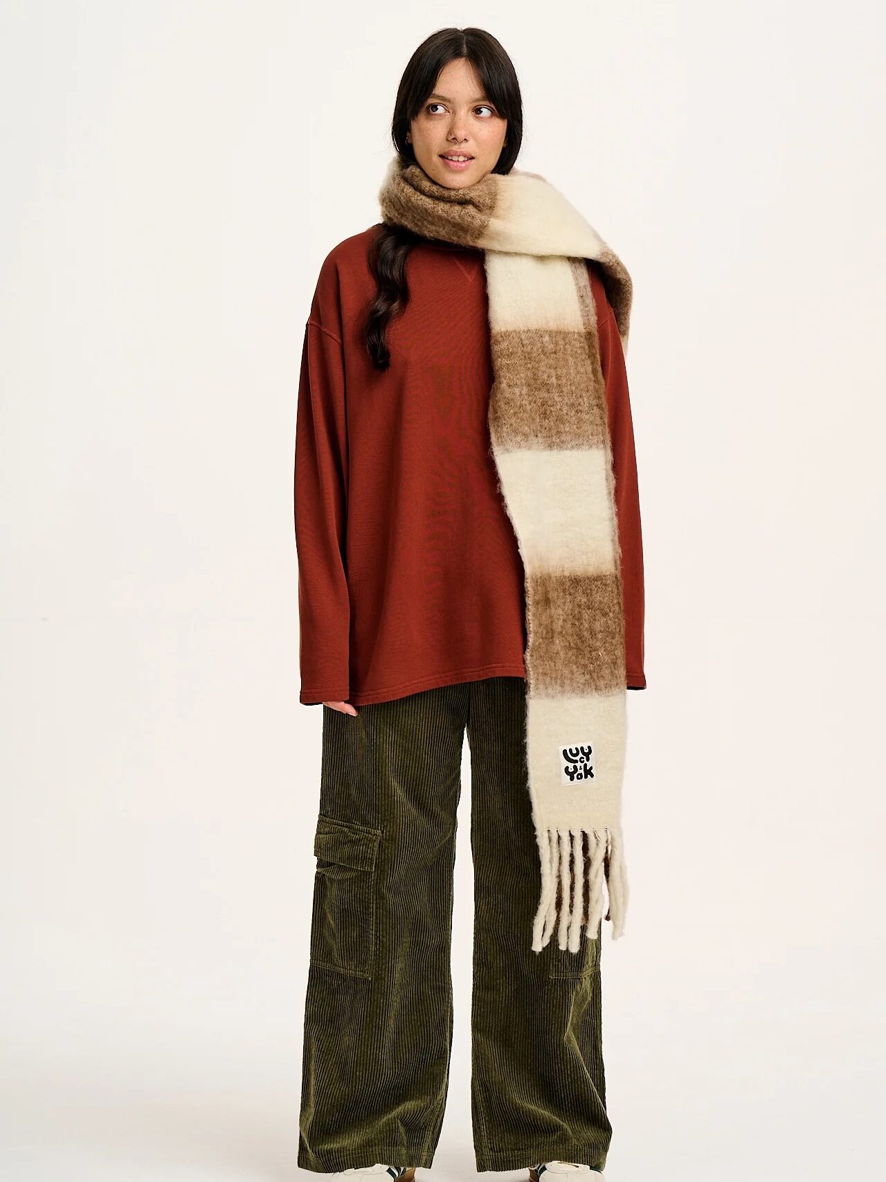 A person wearing a brown sweater, green corduroy pants, and a long plaid scarf stands against a plain background.