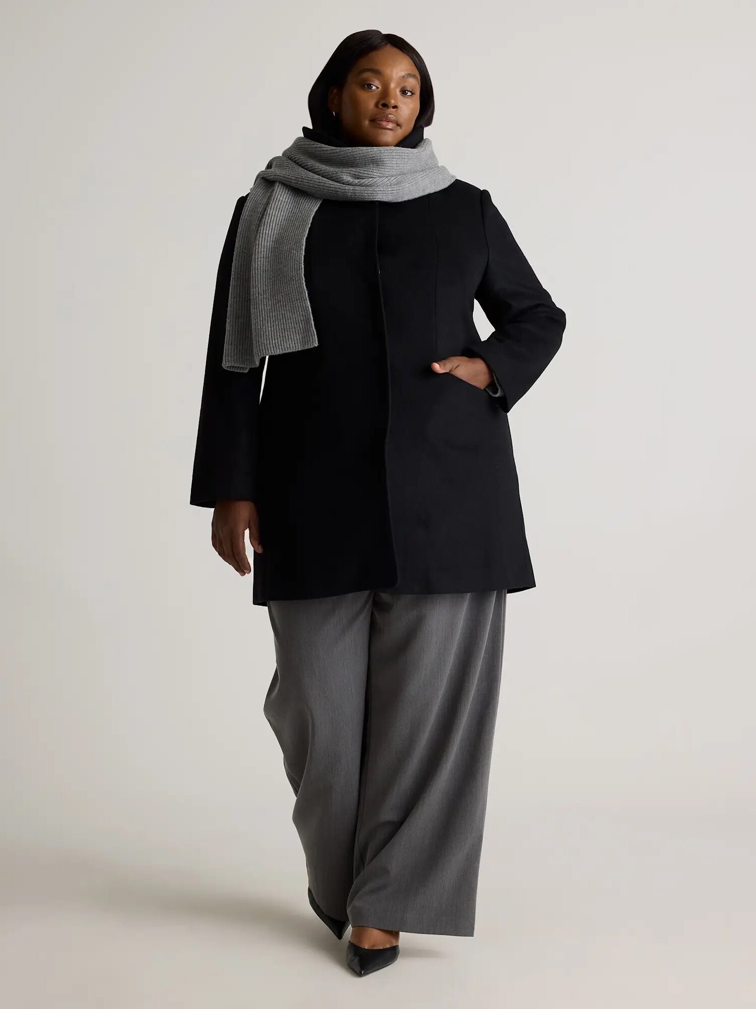 Person wearing a black coat, grey scarf, and grey wide-legged pants, walking confidently against a plain background.