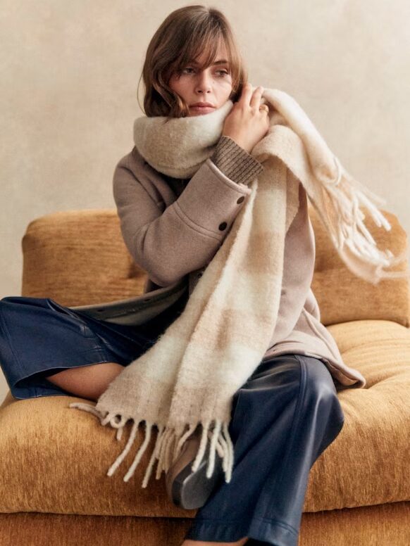 Person sitting on a mustard-colored chair, wearing a beige coat and a large plaid scarf.