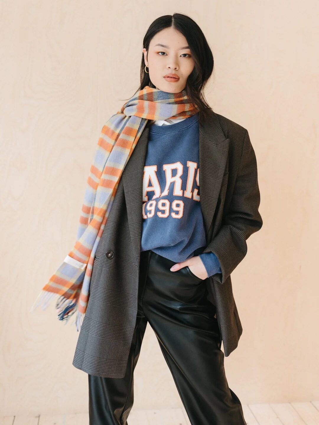 Person wearing a plaid scarf, oversized blazer, blue "Paris 1999" sweatshirt, and black leather pants stands against a plain background.